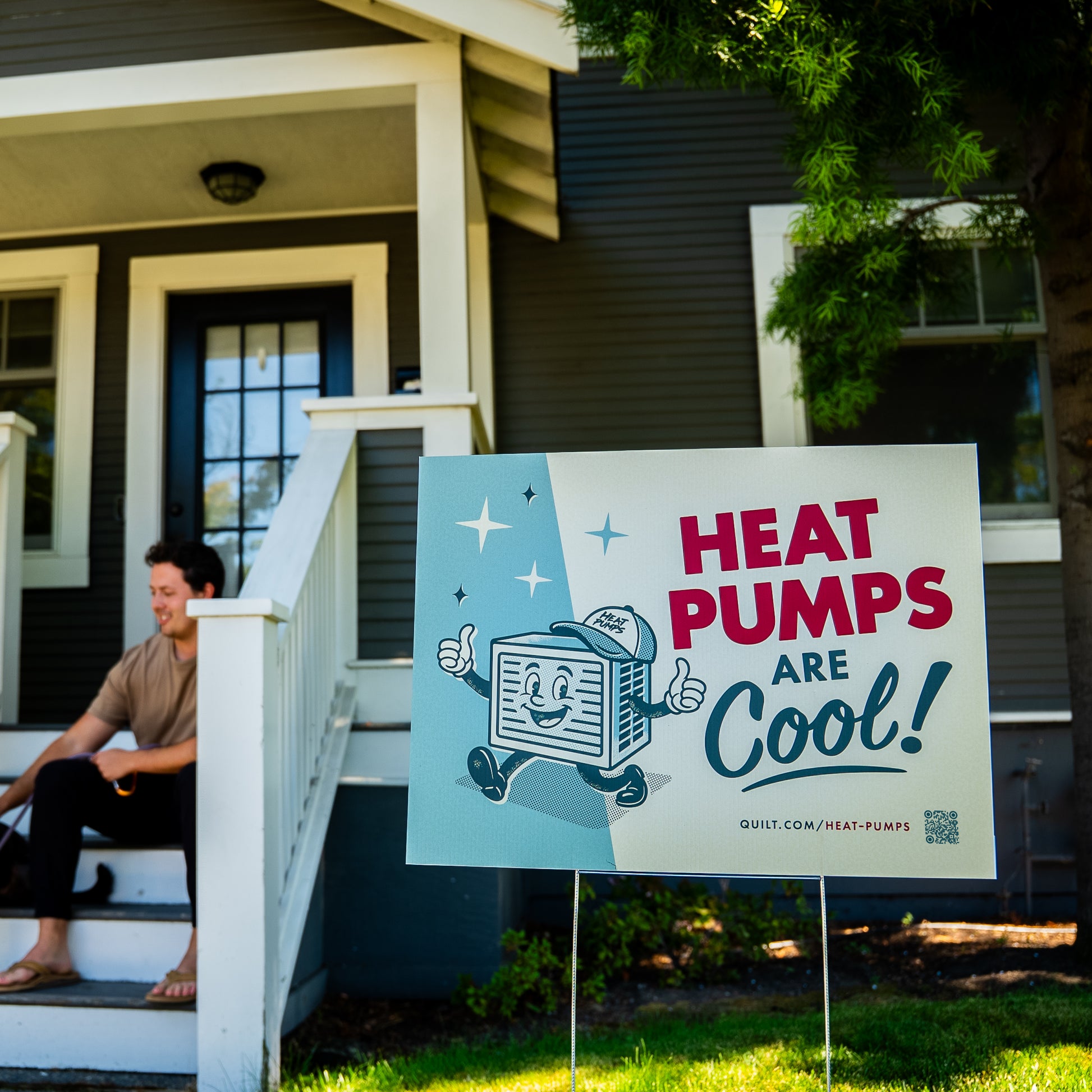 Person sitting on a house porch with a 'Heat Pumps Are Cool!' sign in front.
